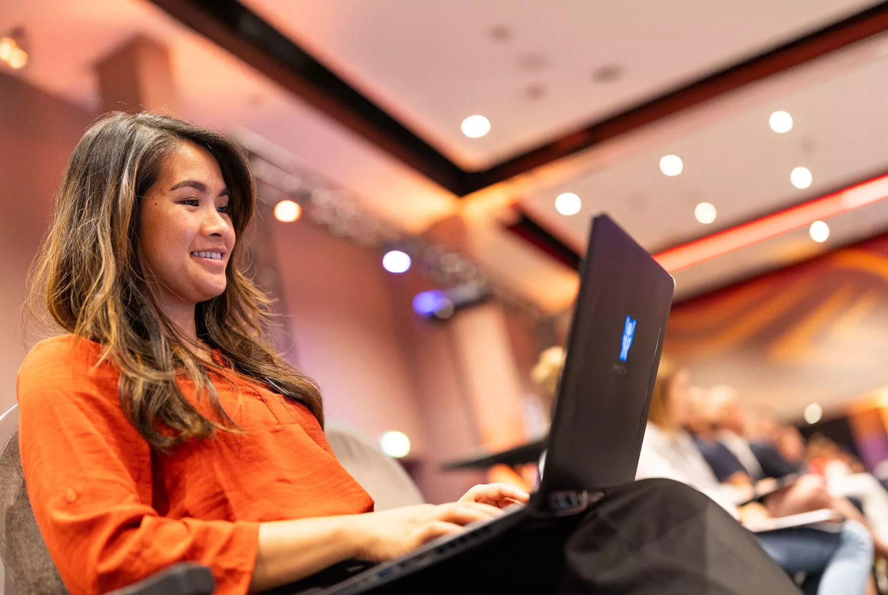 Vrouw zit in een zaal met haar laptop op schoot en lacht naar het scherm